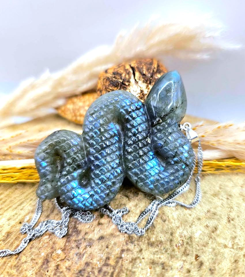 A snake-shaped pendant with a textured, scale-like pattern and an iridescent blue-green labradorite finish, shown on a silver chain with dried grass and a small wooden piece as props.