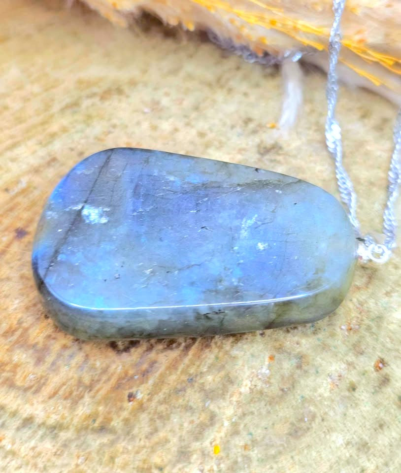 A polished labradorite stone pendant necklace with a silver chain, resting on a textured tan surface.