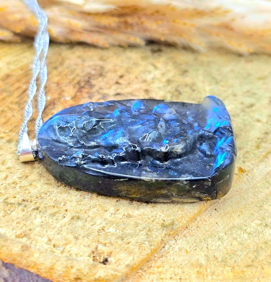 A carved black stone pendant with vibrant blue iridescent opal inclusions, suspended from a silver cord, resting on a textured tan stone surface.
