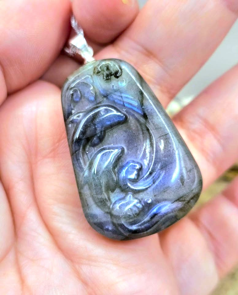A hand holding a carved labradorite pendant with a dolphin design. The pendant displays a natural play-of-color with gray, blue, and purple flashes, suspended from a silver-colored chain.