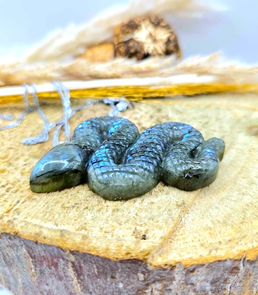 A small coiled iridescent green bead or charm with a glossy finish, resting on a textured tan surface with dried natural elements in the background.