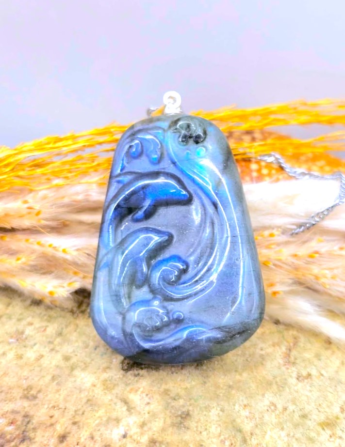 A carved blue stone pendant with a dolphin design, hanging from a silver chain, displayed on a textured surface with dried grass.