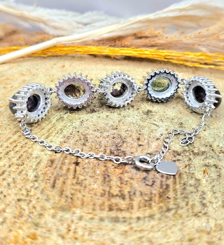 A silver chain bracelet with five circular spiky links, each containing a small inlaid stone in shades of green, pink, and purple, displayed on a textured wood slice with dried grass.