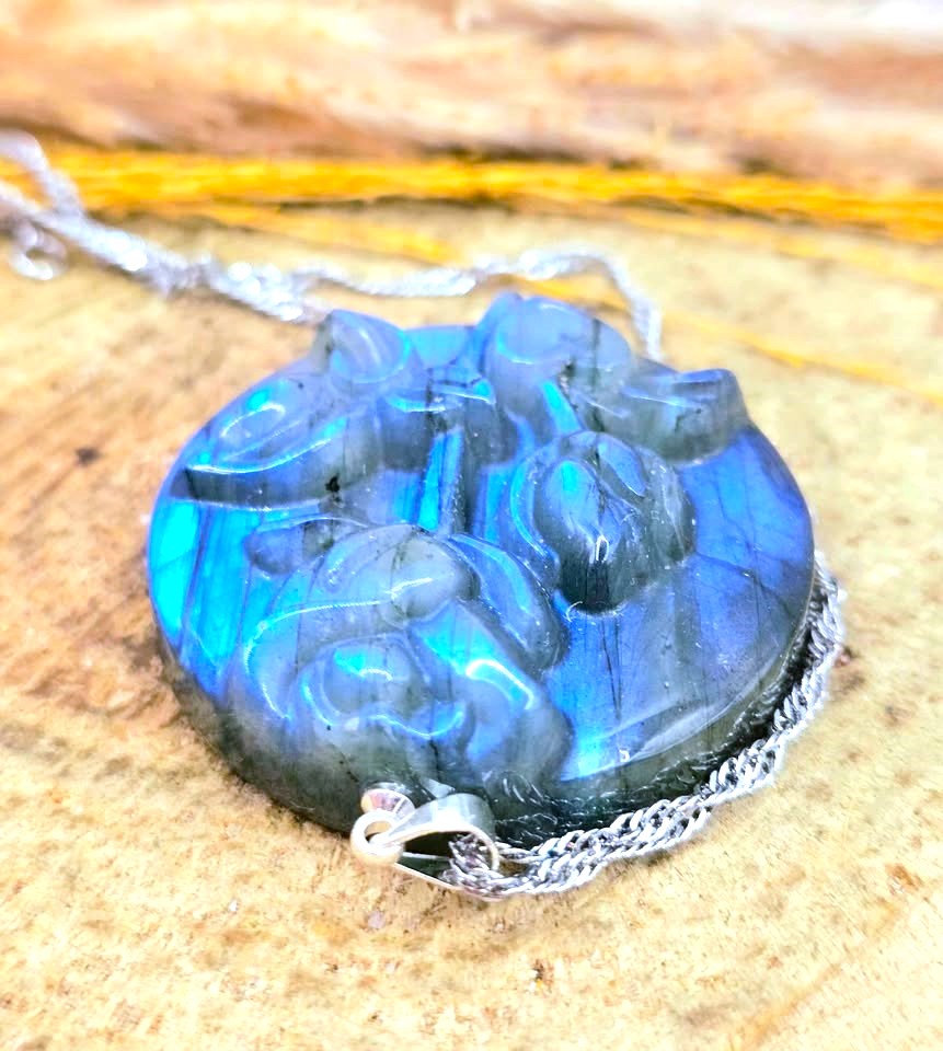 A carved labradorite pendant necklace with a silver chain, resting on a textured wood surface. The pendant displays vibrant blue and green iridescent labradorite with dark gray matrix lines.