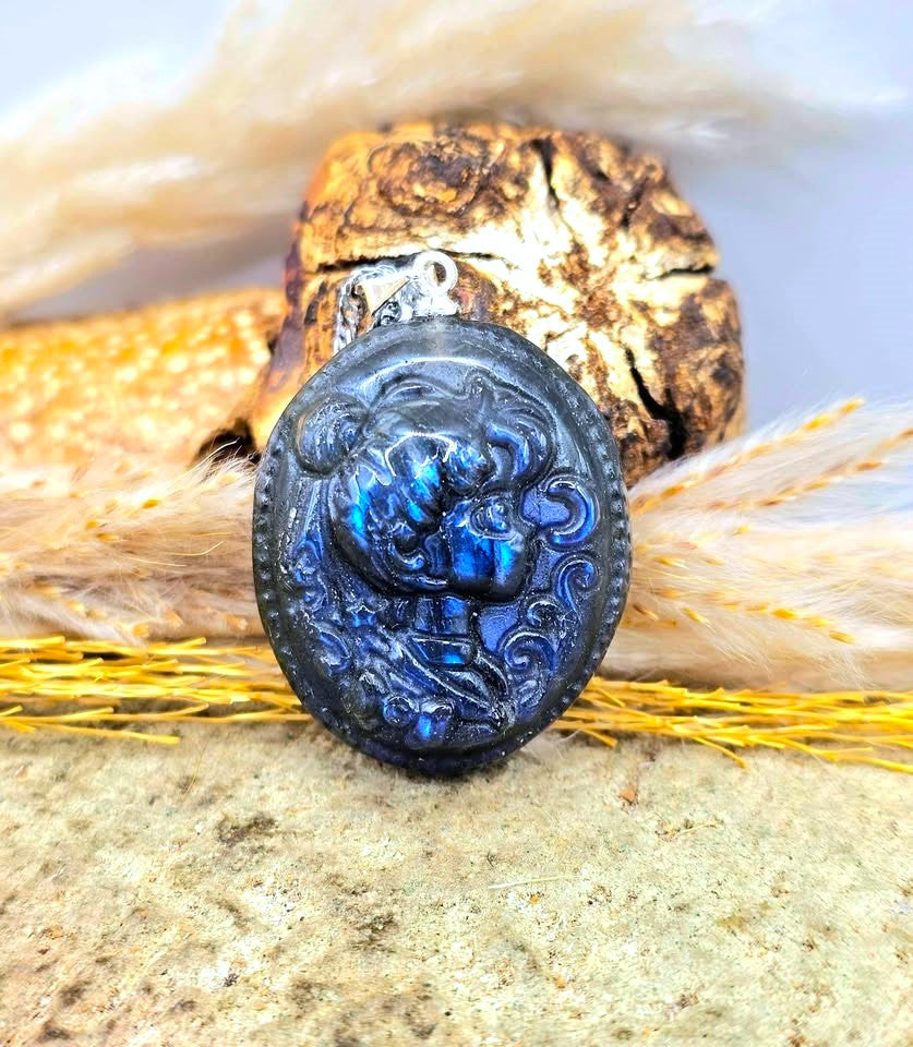 A round dark blue and black carved pendant with an embossed floral or creature design, resting on natural dried plant material and a stone surface.