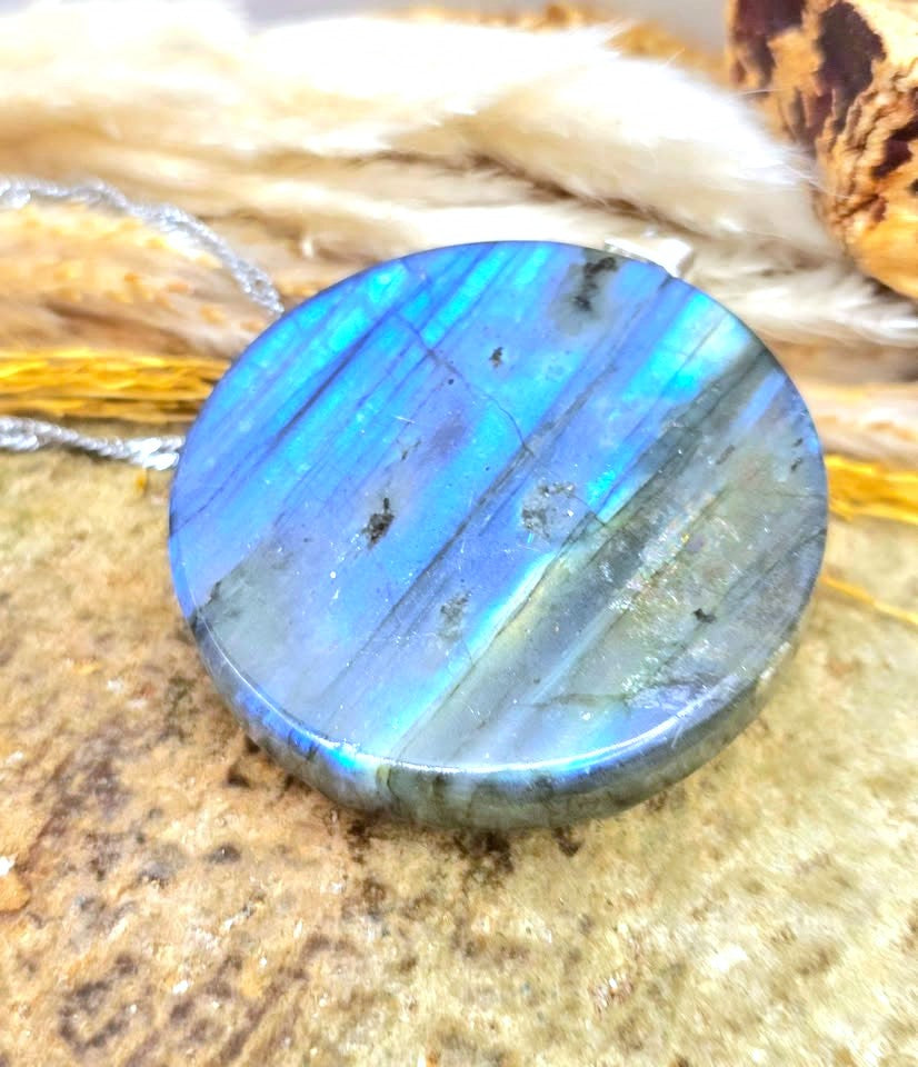 A round labradorite gemstone cabochon pendant with vibrant blue and green flashes, resting on a textured stone surface with dried grass in the background.