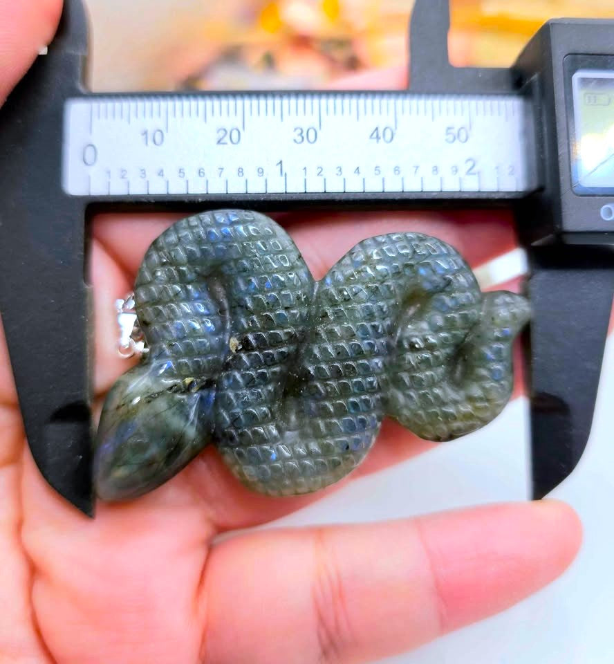 A hand holds a carved snake-shaped object under a digital caliper to show its size. The item appears to be a dark gray to black stone or resin carving with a textured scale pattern.
