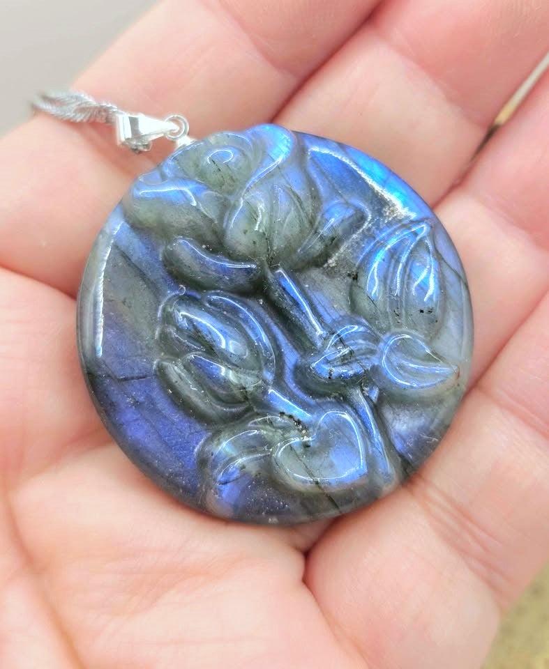A round labradorite pendant with a carved floral relief, held in a person's hand. The pendant displays a blue-gray color with a strong iridescent blue flash.