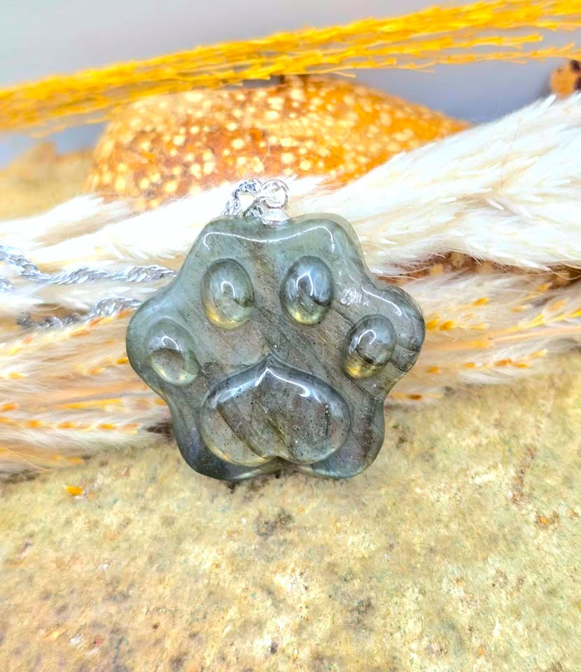 A paw print pendant necklace made of polished dark gray stone, resting on a textured surface with white and orange feathers in the background.