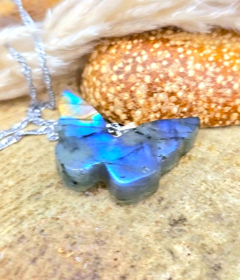 A close-up of a silver necklace with a butterfly-shaped pendant made of iridescent labradorite stone, resting on a sandy surface with a starfish and white feathers in the background.