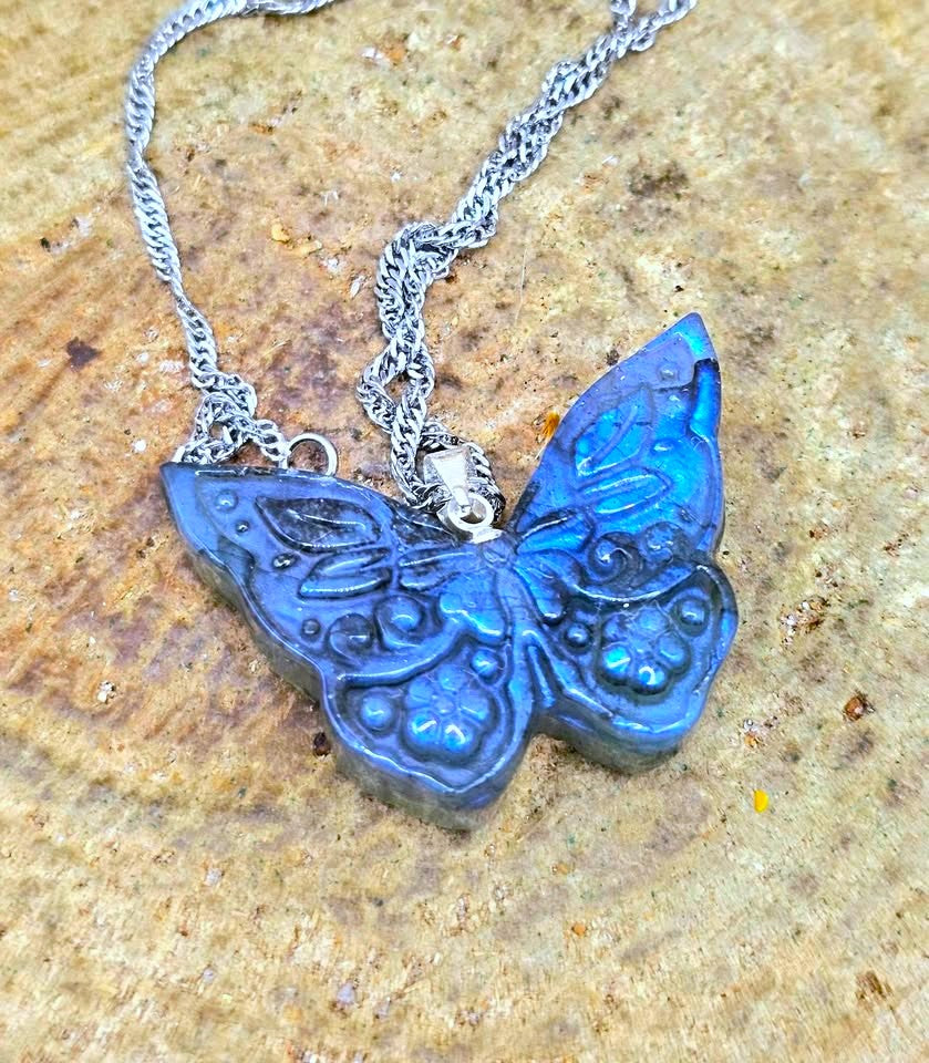 A butterfly-shaped pendant necklace with a textured blue body and a silver chain, displayed on a textured tan stone surface.