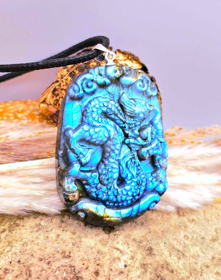 A carved blue iridescent dragon pendant on a black cord, displayed on a textured surface with fur and a small piece of wood.