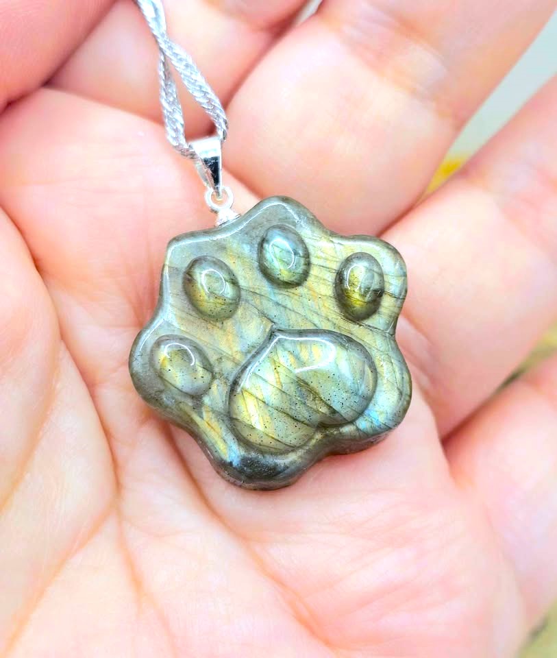 A close-up of a silver paw-shaped pendant with a greenish metallic, iridescent stone inlay, held in a person's hand.