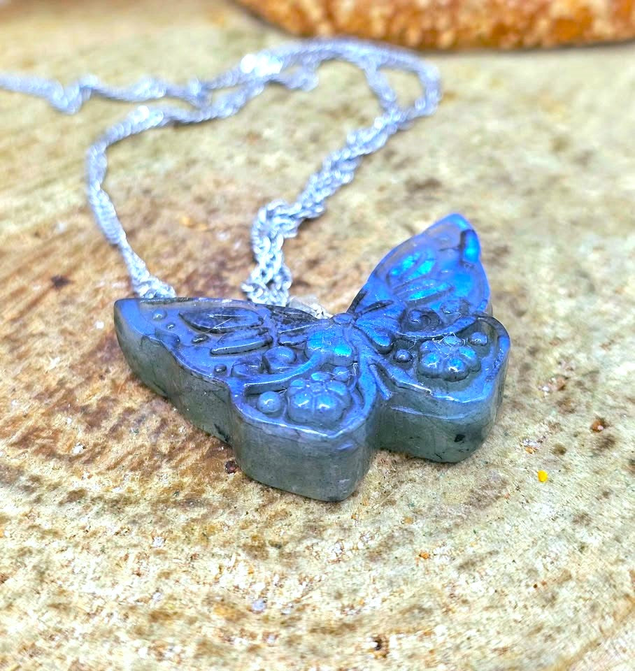 A butterfly-shaped pendant necklace made of dark blue-gray stone with iridescent blue highlights, hanging from a silver chain on a textured sandy surface.