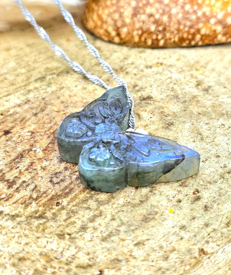 A silver necklace with a butterfly-shaped pendant made of polished labradorite, resting on a textured stone surface.