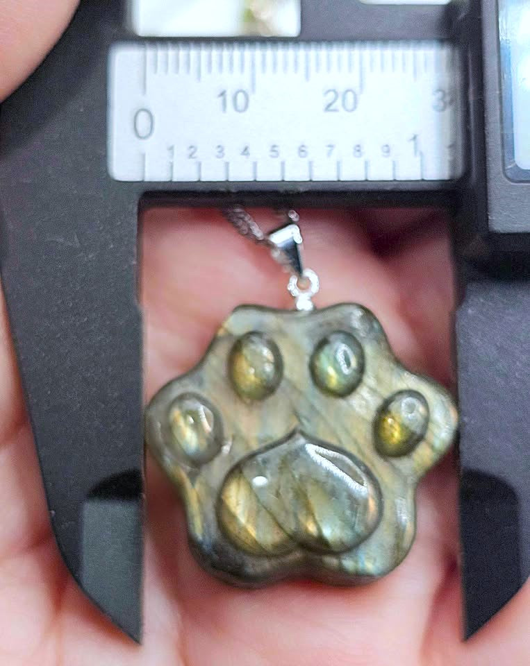 A paw-shaped pendant made of polished labradorite stone being measured under a caliper, held in a hand. The pendant has a silver-tone loop at the top and displays natural labradorite chatoyancy with olive green, gold, and gray tones.