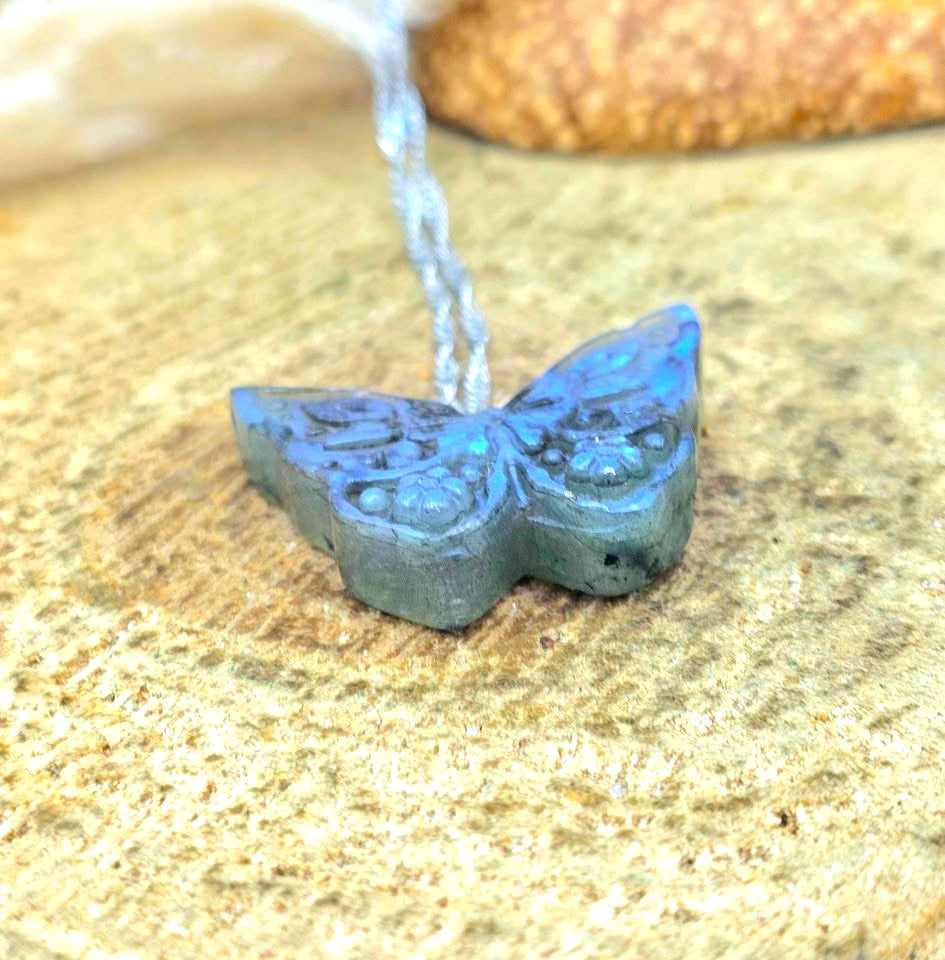 A carved butterfly-shaped pendant in a blue-gray stone with a light blue cord, resting on a textured sandy surface.