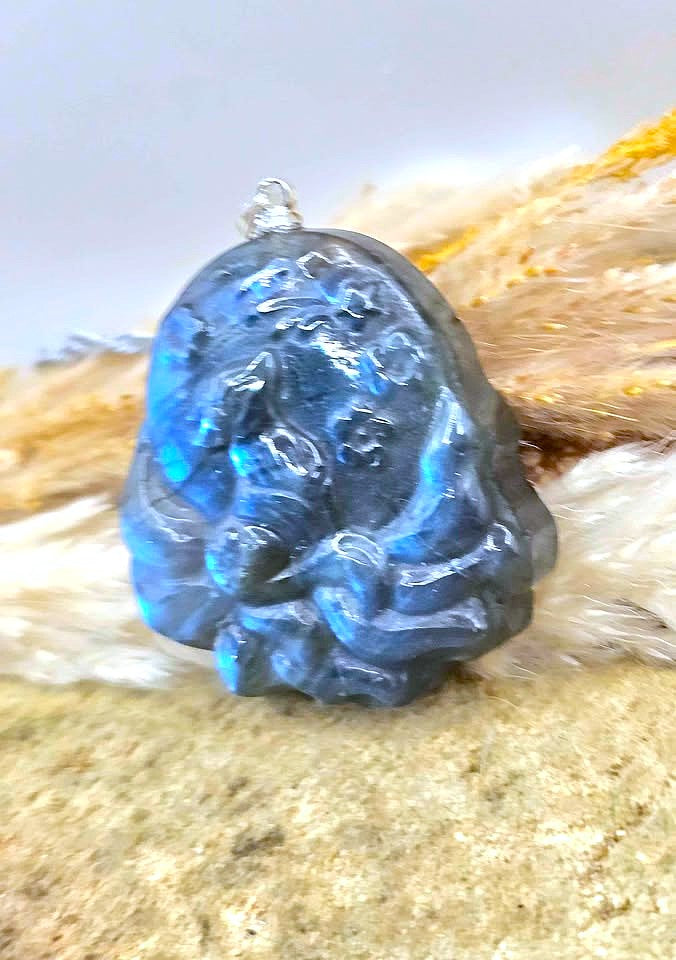 A carved blue labradorite nine tails fox pendant with a small silver bail, resting on a textured beige surface with soft white and tan fur in the background.