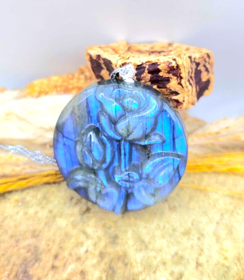 A round cabochon pendant featuring a carved floral motif in vibrant blue labradorite with green and gray iridescent flashes, resting on a textured stone surface.