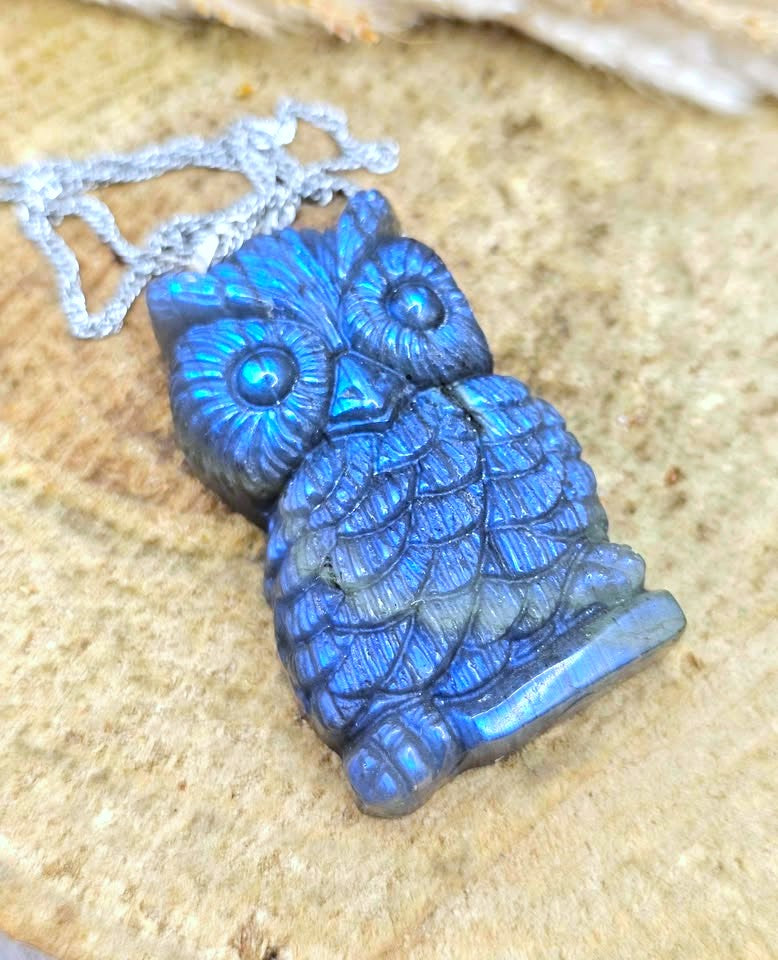 A carved blue owl pendant with an iridescent labradorite finish, hanging from a silver-toned chain against a textured beige stone background.