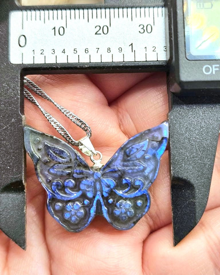 A close-up of a butterfly-shaped pendant necklace being measured by a caliper over a hand. The pendant is dark blue with lighter blue floral details and a silver-toned chain.