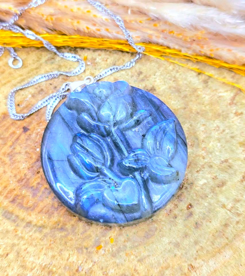 A round carved pendant necklace made of blue-gray stone with floral relief details, hanging from a silver-toned chain on a textured tan surface with dried grass in the background.