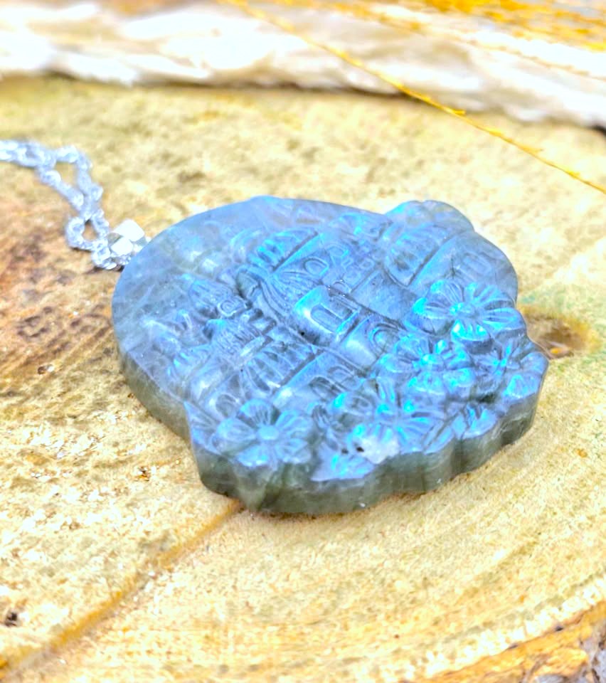 A carved blue labradorite pendant necklace with a silver chain, resting on a textured natural stone surface.