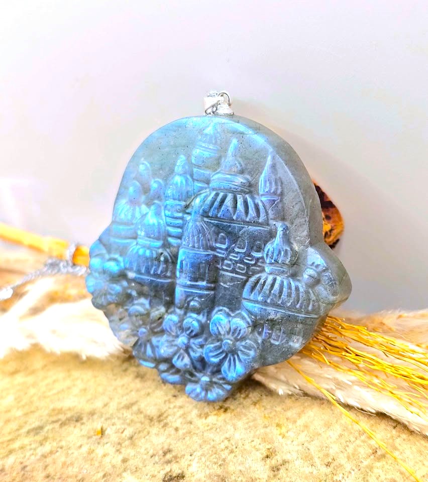 A blue-gray carved stone pendant featuring a detailed architectural design with domes and floral motifs at the base, shown resting on a textured beige surface with dried grass.