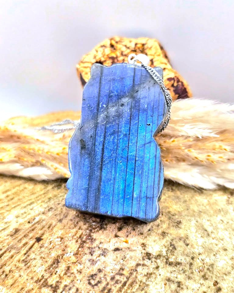 A blue labradorite stone pendant necklace with a silver chain, displayed on a textured stone surface with dried grass and a rock in the background.