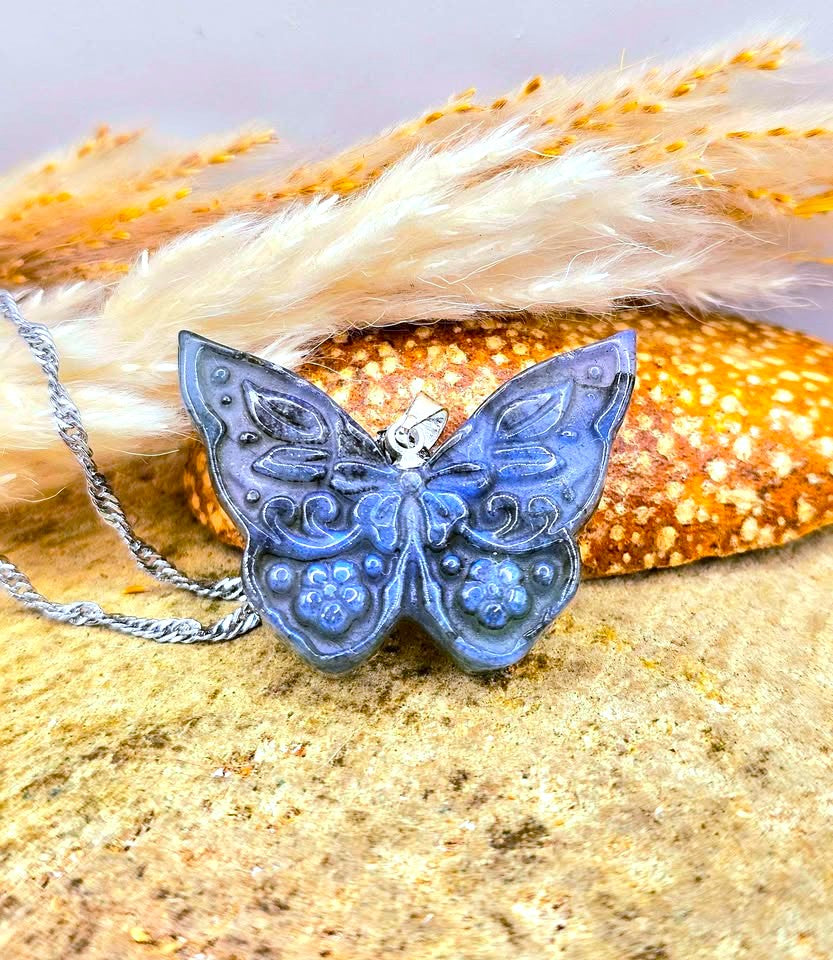 A blue butterfly pendant necklace with a silver chain, displayed on a sandy surface with dried grass and a small orange flower.