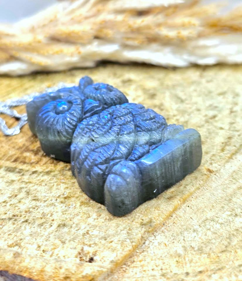 A carved labradorite owl pendant with a silver chain, resting on a textured sandy surface with a blurred natural background.