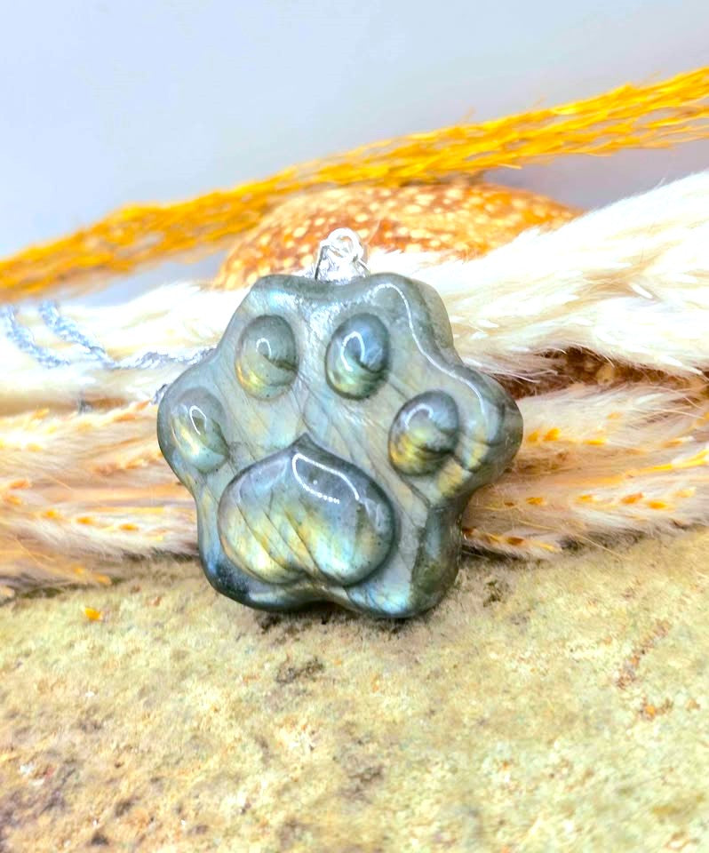 A paw-shaped pendant made of iridescent labradorite stone, resting on a textured surface with white and orange feathers in the background.