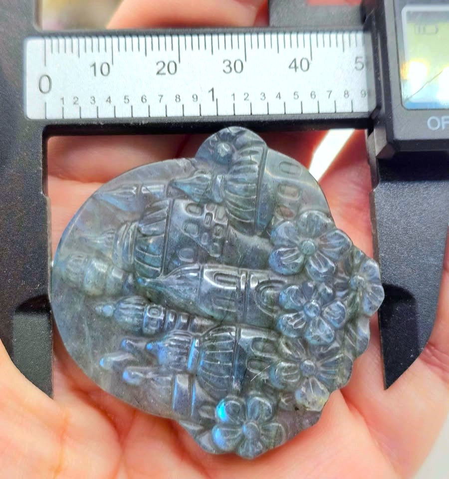 A close-up of a hand holding a carved labradorite pendant under a digital caliper to measure its size. The pendant features a detailed relief carving of a castle and flowers, showing a blue-gray iridescent labradorite finish.
