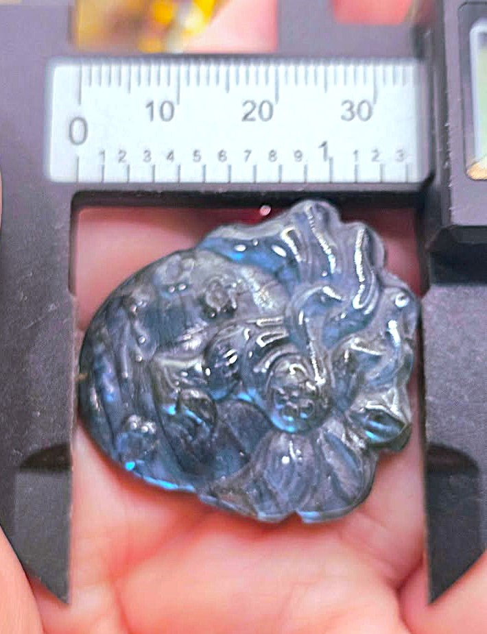A close-up of a carved dark blue-gray stone pendant held in a hand under a caliper with a ruler scale visible above it, showing its size.