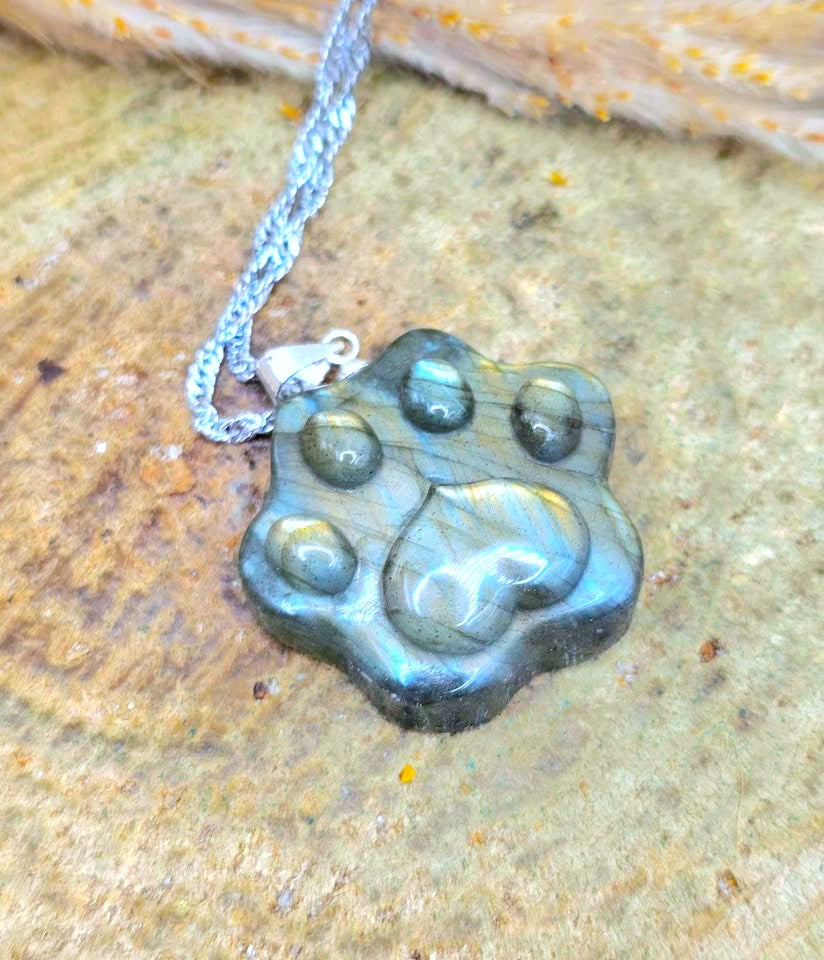 A paw-shaped pendant necklace made of polished labradorite stone with a silver chain, resting on a textured natural rock surface.