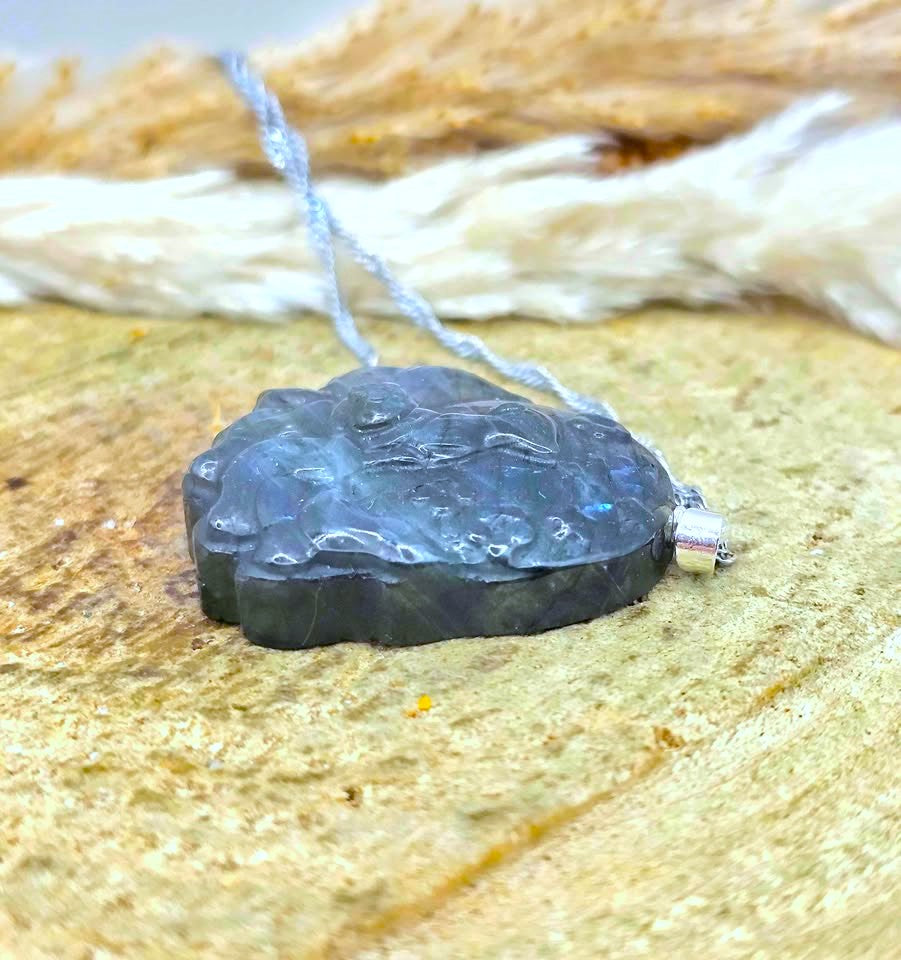 A carved dark blue-gray labradorite pendant necklace with a silver bail, resting on a textured sandy surface with a blurred natural background.