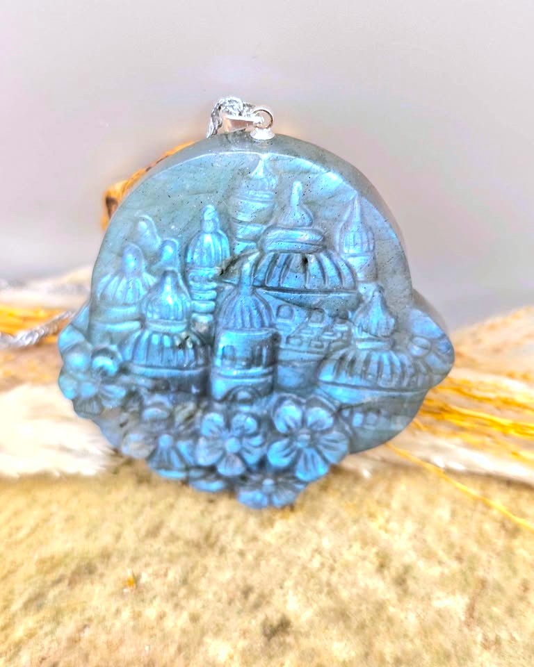 A round blue carved pendant featuring an embossed design of domed buildings and flowers, with a small silver loop at the top, displayed on a textured surface.