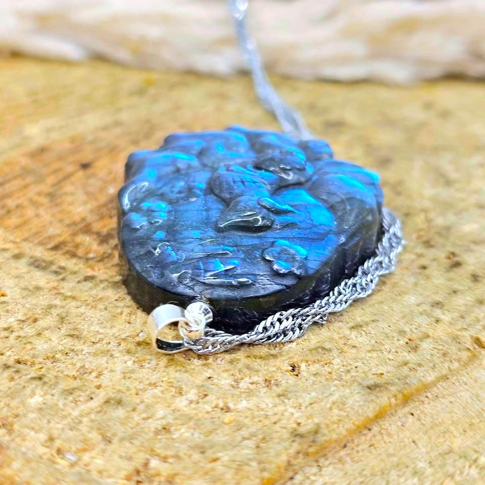 A close-up of a carved black stone pendant with vibrant blue iridescent inclusions, attached to a silver chain and resting on a textured tan stone surface.
