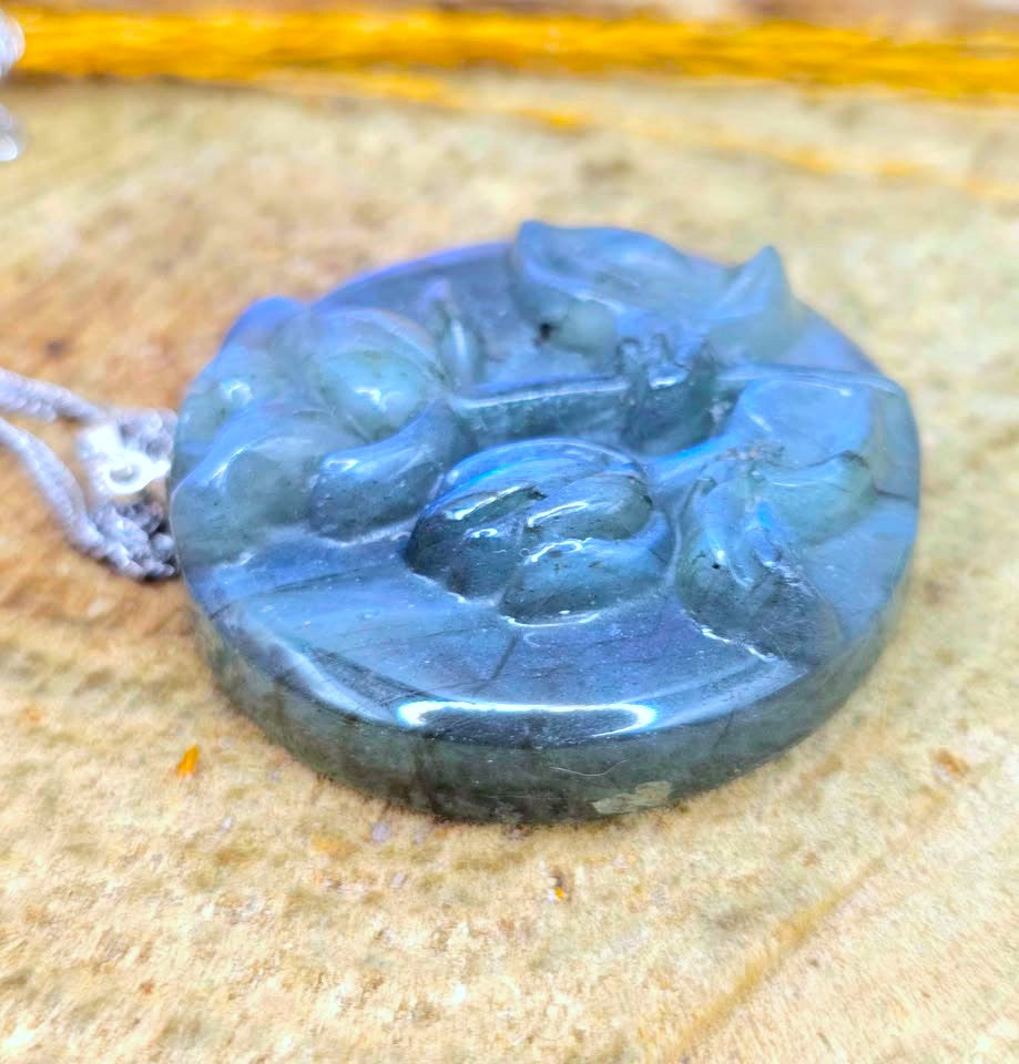 A carved round labradorite pendant with a blue-gray sheen, resting on a textured tan stone surface with a silver-colored cord visible on the left.