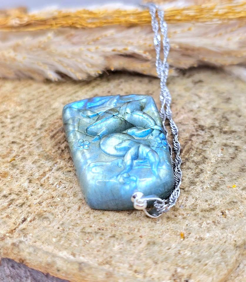 A rectangular pendant necklace with a carved figure design, featuring a blue-green iridescent finish and a silver chain, displayed on a textured stone surface.