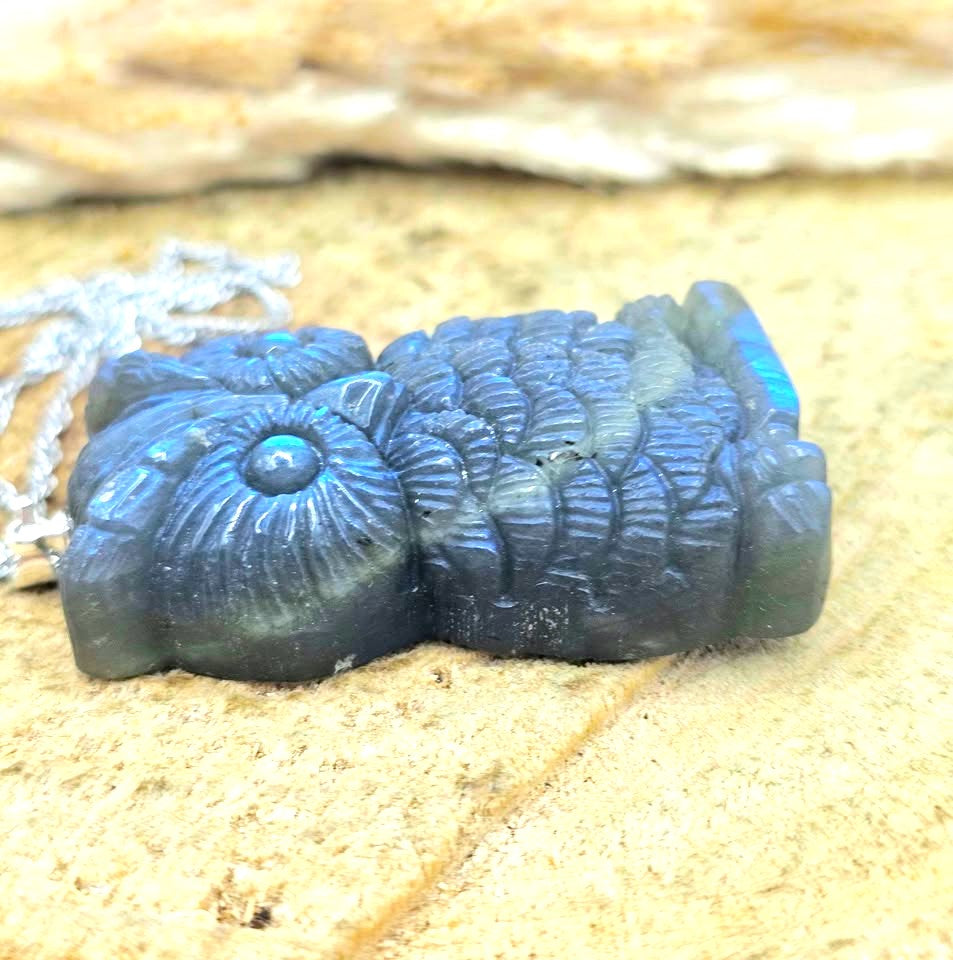 A carved blue-gray stone owl pendant with a silver chain, resting on a sandy surface with a blurred natural background.