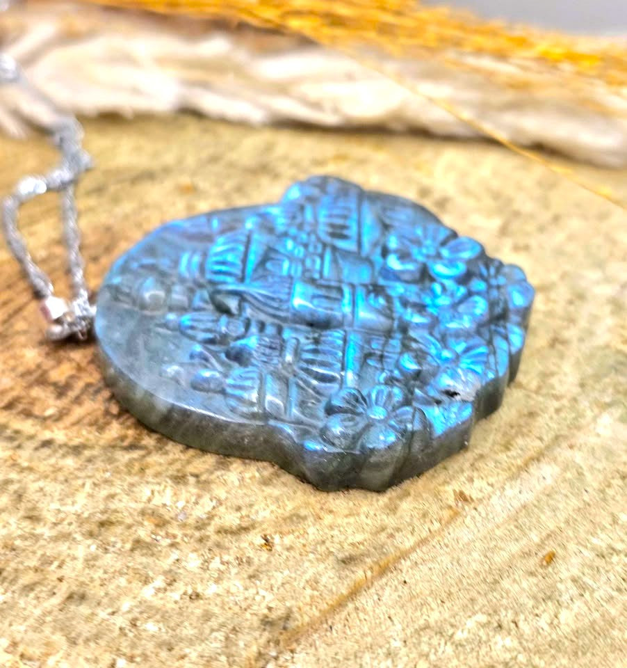A carved labradorite pendant necklace with a silver chain, resting on a textured stone surface. The pendant features a natural blue flash characteristic of labradorite.