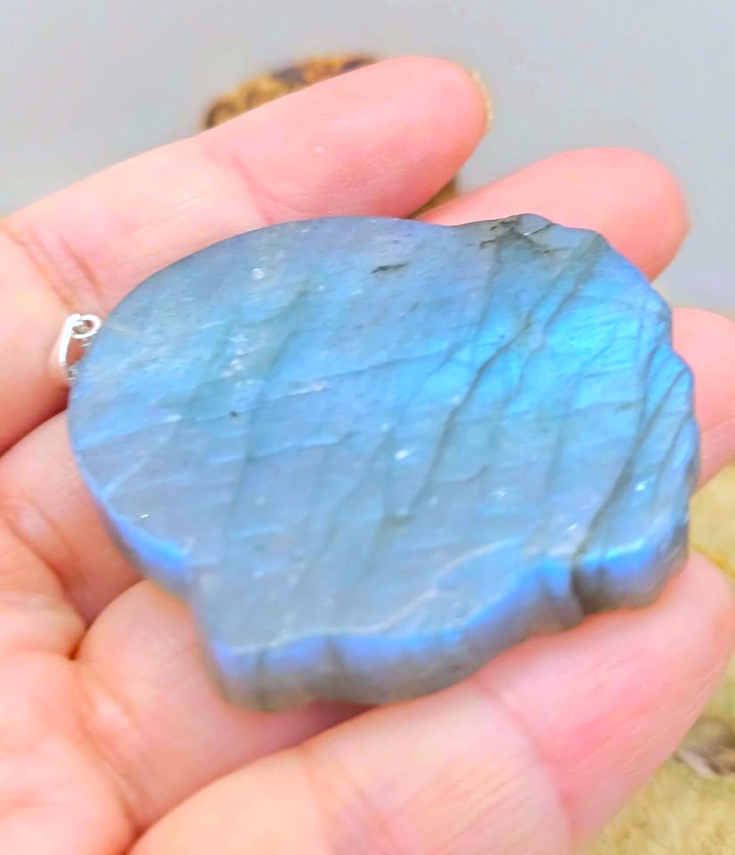A hand holding a polished irregular-shaped labradorite stone pendant with a shimmering blue iridescent sheen and subtle gray veining.