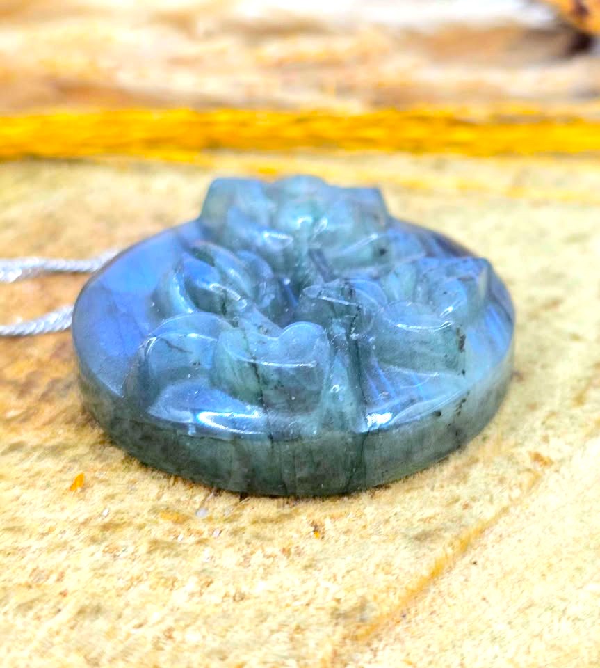 A round carved labradorite pendant with a floral relief design, resting on a textured sandy surface with a blurred natural background.