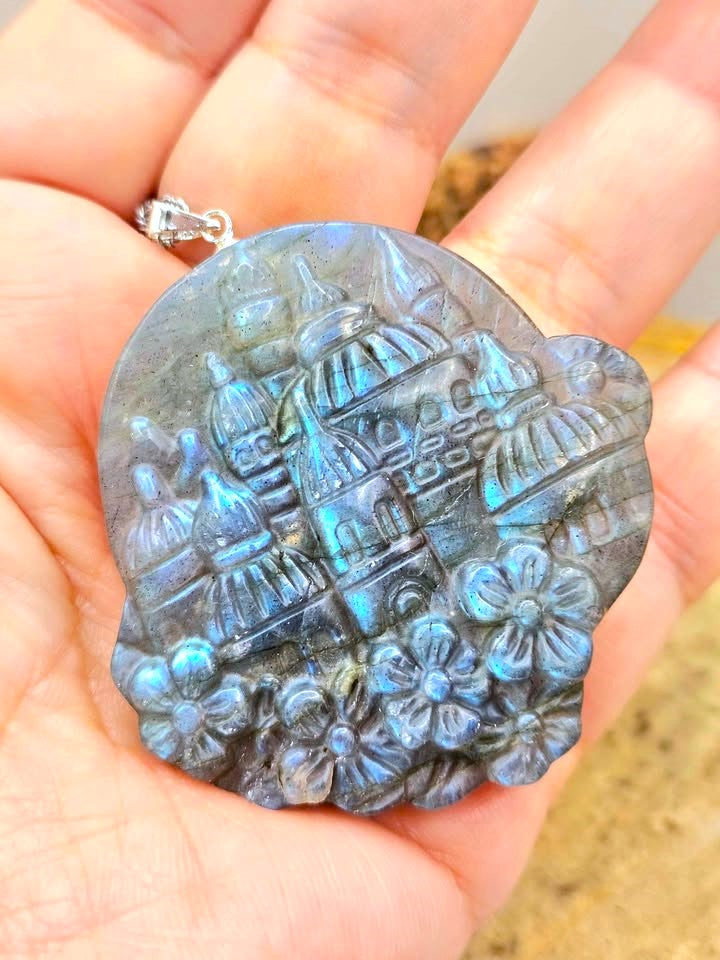 A hand holding a round labradorite pendant featuring a raised relief of a domed palace and floral motifs, showing a blue iridescent sheen.