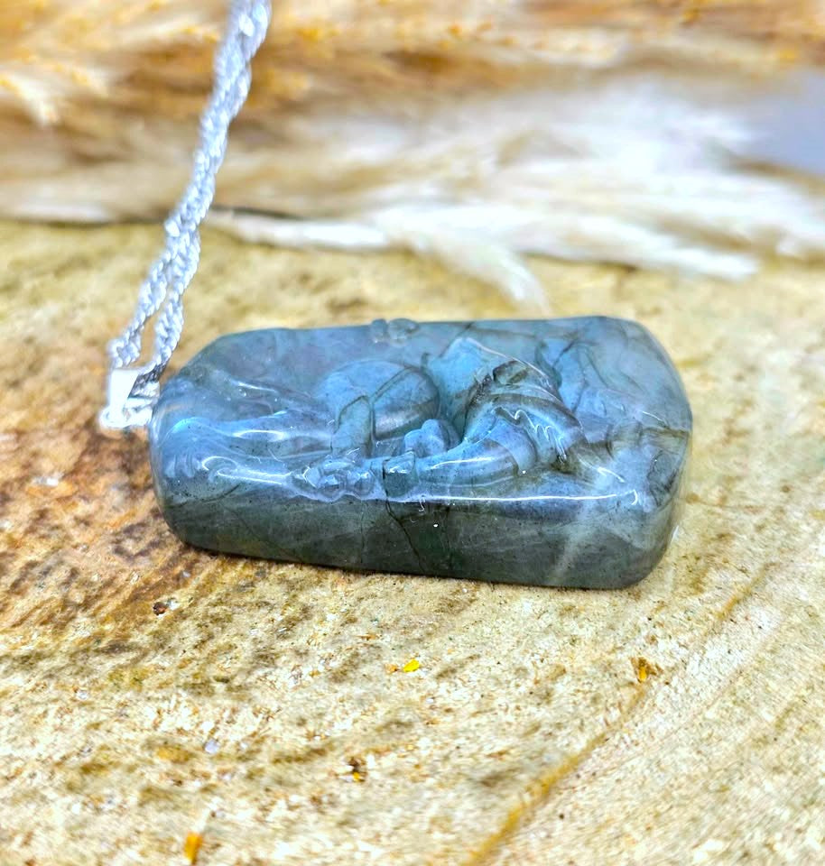 A carved Labradorite pendant necklace with a silver-toned chain, resting on a textured natural stone surface.