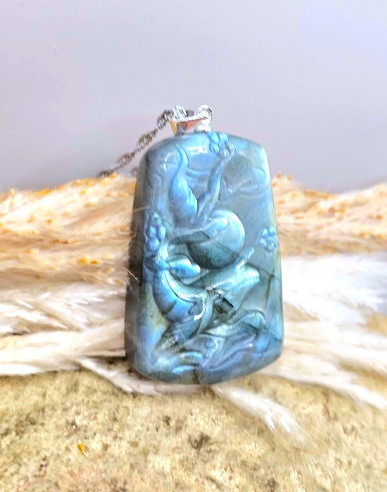 A carved labradorite pendant necklace with a blue-green sheen and dark inclusions, resting on a textured beige surface with white faux fur.