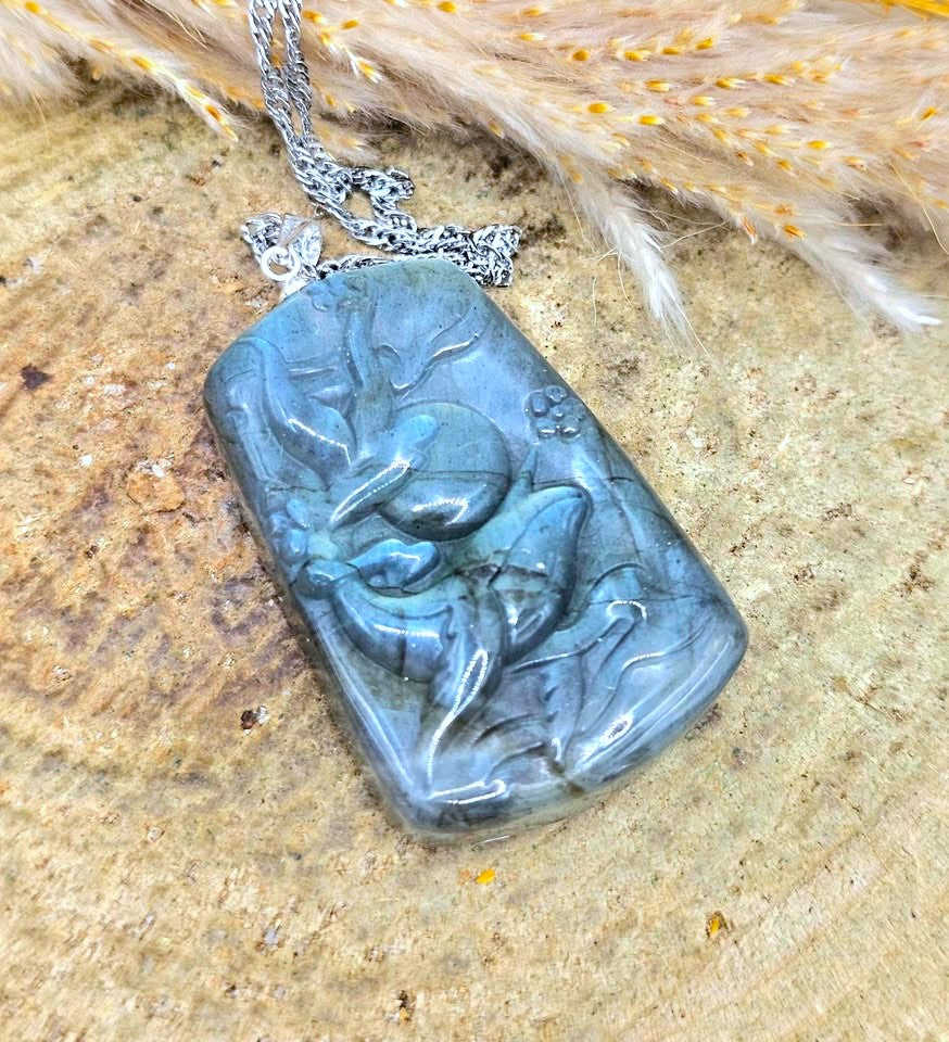 A carved rectangular labradorite pendant necklace with a silver chain, displayed on a textured stone surface with dried grass.