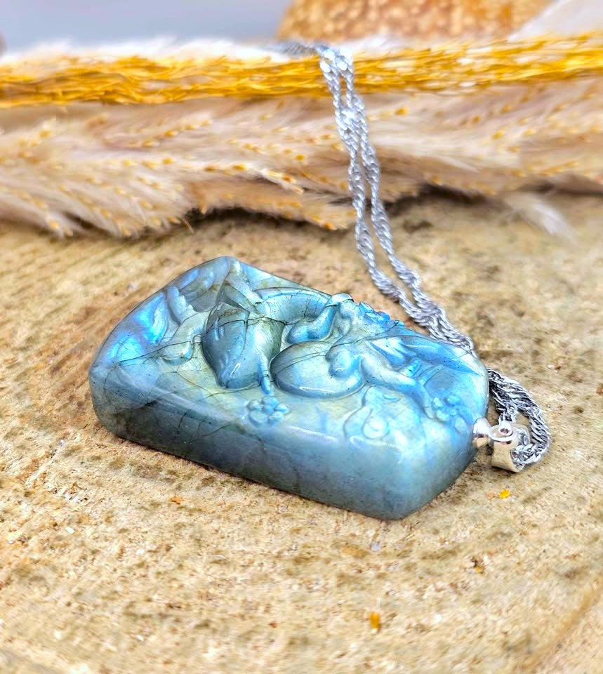 A carved labradorite pendant necklace with a silver chain, resting on a textured sandy surface with dried grass in the background.