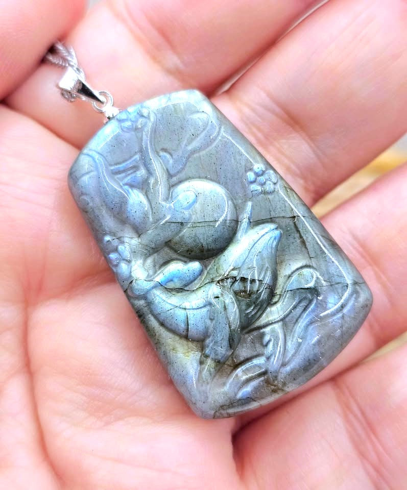 A rectangular labradorite pendant with a carved dragon design, held in a person's hand. The pendant exhibits a gray-blue color shift with metallic silver highlights.